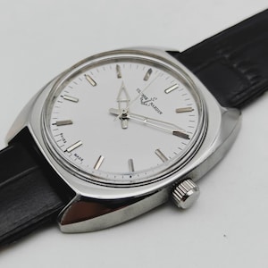 May include: A silver wristwatch with a white face and a black leather strap. The watch face features silver hour markers and hands. The brand name "ULYSSE NARDIN" is printed on the watch face. The watch has a classic, elegant design.