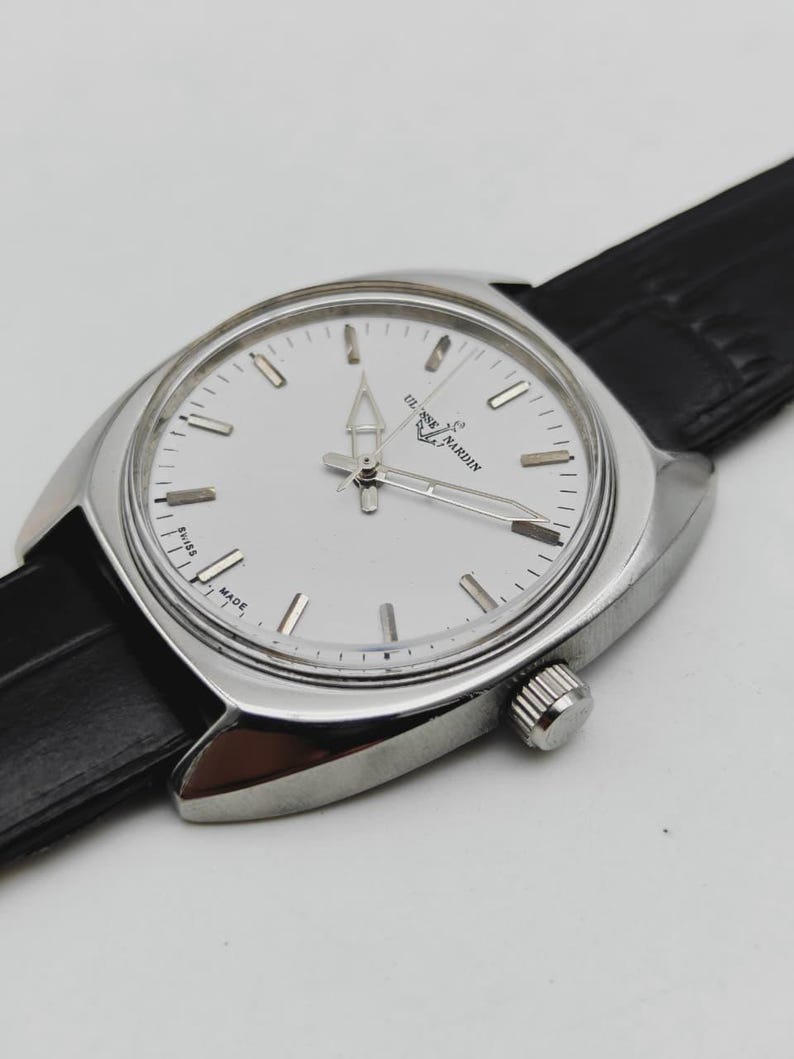 May include: A silver wristwatch with a white face and a black leather strap. The watch face features silver hour markers and hands. The brand name "ULYSSE NARDIN" is printed on the watch face. The watch has a classic, elegant design.