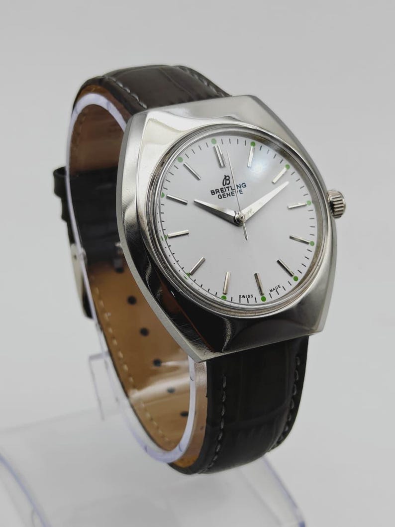 May include: A Breitling Geneve watch with a silver-toned case and a white dial. The watch features a black leather band and a silver crown. The dial has silver hands and green markers. The watch is a classic timepiece.