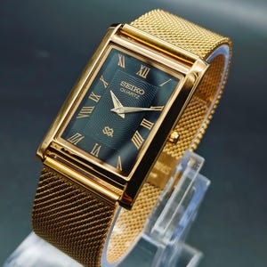 May include: A gold-toned Seiko Quartz watch with a rectangular face and mesh band. The watch face is dark green with gold Roman numerals and hands. The watch is displayed on a clear stand.