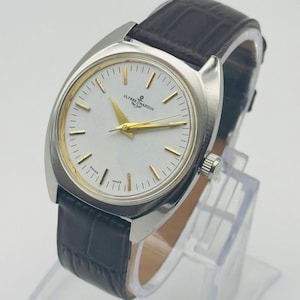 May include: A silver-toned Ulysse Nardin wristwatch with a white face and gold hands and markers. The watch has a dark brown leather band. The watch is displayed on a clear stand.