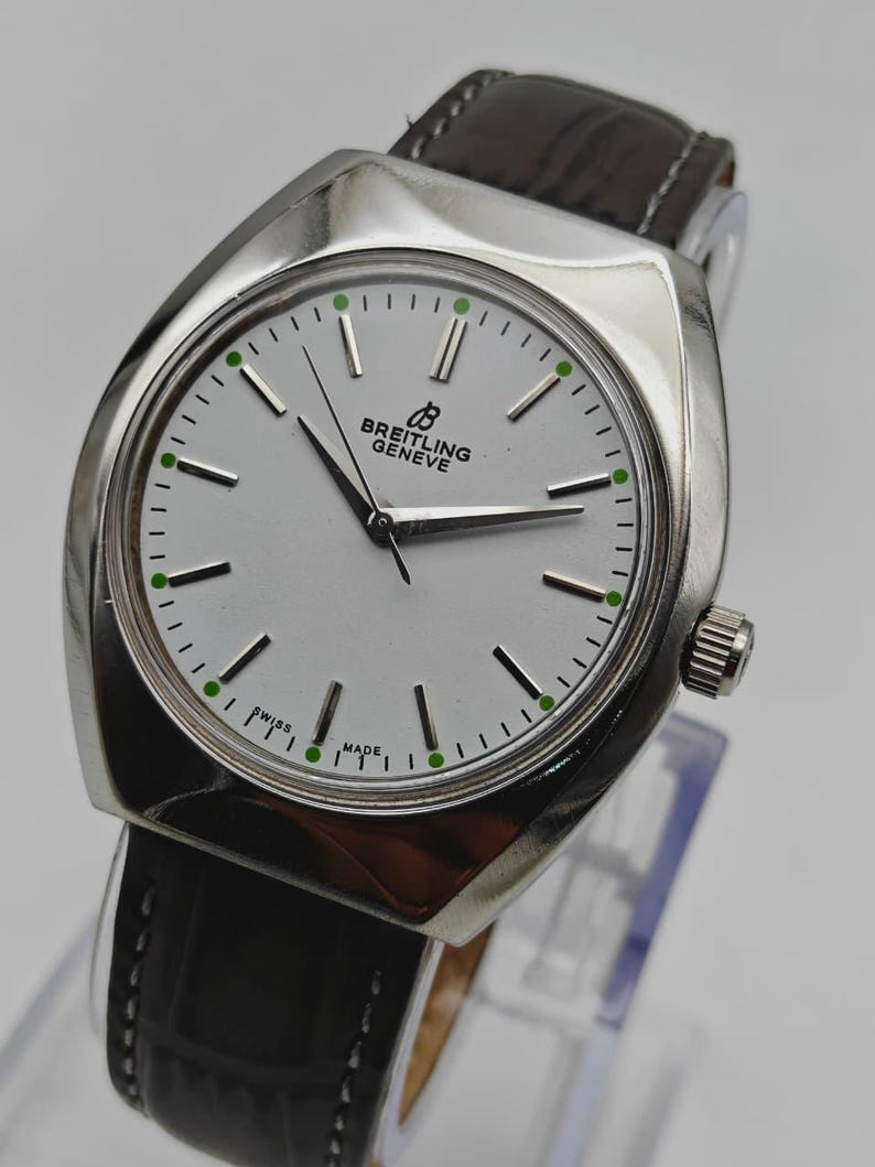 May include: A silver-toned Breitling Geneve watch with a white face and black hands. The watch has a black leather band and green accents on the face. The watch face reads "SWISS MADE".