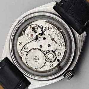 May include: A detailed view of a vintage Ulysse Nardin watch, revealing its mechanical workings through a transparent back. The watch has a silver case, black leather strap, and the text "SWISS SEVENTEEN 17 JEWELS ULYSSE NARDIN".