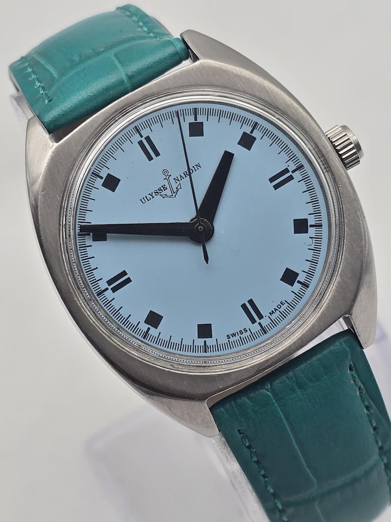 Pu&ograve; includere: Un orologio Ulysse Nardin vintage con quadrante azzurro e lancette e indici neri. L'orologio ha una cassa argentata e un cinturino in pelle verde acqua. Il quadrante mostra il logo Ulysse Nardin e la scritta "SWISS MADE".