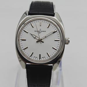 May include: A silver Ulysse Nardin wristwatch with a white face and black leather strap. The watch has a round face with silver hands and markers. The watch is marked "Swiss Made".