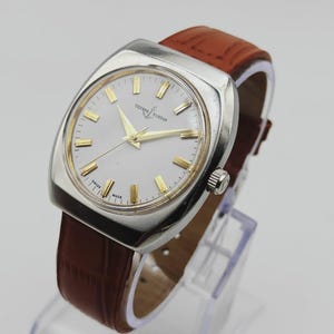 K&ouml;nnte beinhalten: Eine Vintage-Armbanduhr von Ulysse Nardin mit silberfarbenem Zifferblatt und goldfarbenen Stundenmarkierungen und Zeigern. Die Uhr hat ein Edelstahlgeh&auml;use und ein braunes Lederarmband. Die Uhr wird auf einem transparenten St&auml;nder pr&auml;sentiert.
