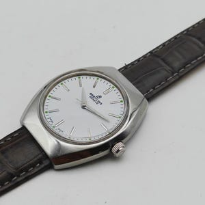 May include: A silver-toned Breitling Geneve watch with a white face and a dark brown leather band. The watch has a rectangular case with a round face and a small crown. The watch face has silver hands and green accents.