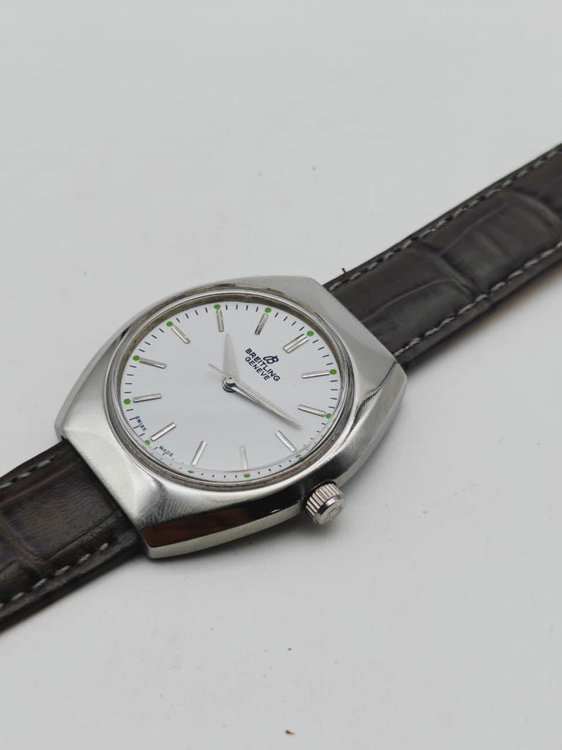 May include: A silver-toned Breitling Geneve watch with a white face and a dark brown leather band. The watch has a rectangular case with a round face and a small crown. The watch face has silver hands and green accents.