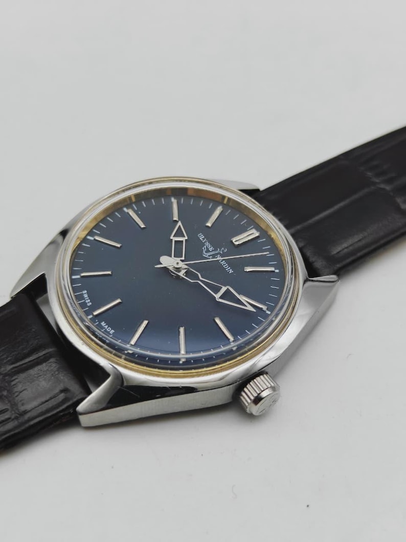 Puede incluir: Un reloj de pulsera plateado con esfera azul oscuro y agujas e indicadores plateados. El reloj tiene una correa de cuero negro. La esfera del reloj tiene la marca "Ulysse Nardin". El reloj es de dise&ntilde;o cl&aacute;sico.