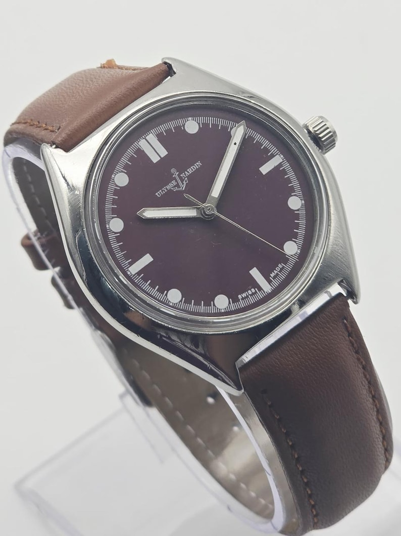 May include: A stainless steel Ulysse Nardin wristwatch with a burgundy dial and brown leather strap. The watch features white hour markers and hands, and a crown on the right side. The watch is marked "SWISS MADE".