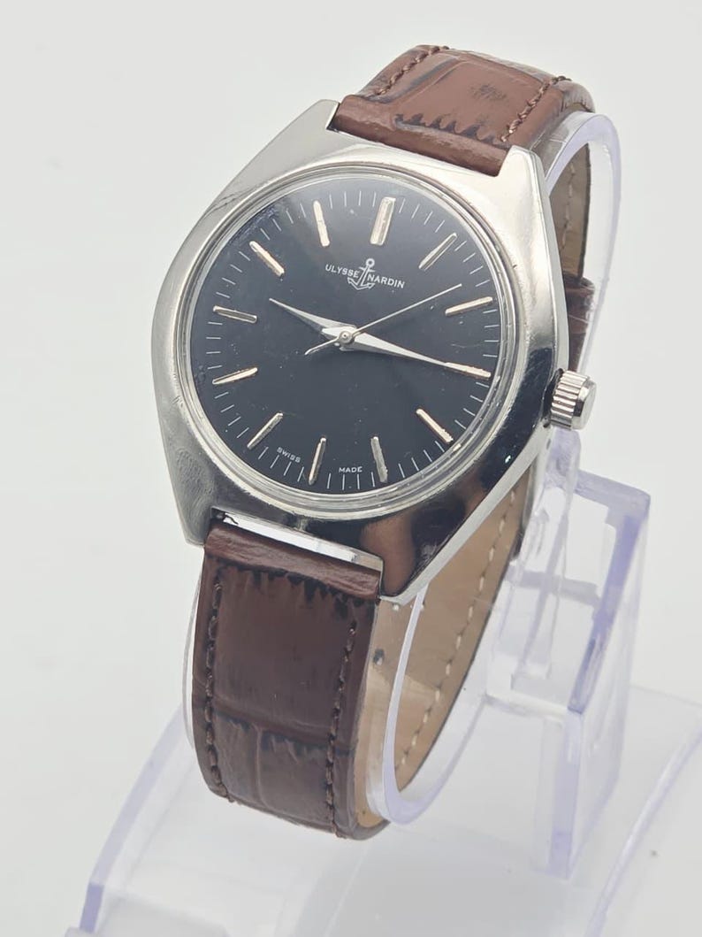 May include: A vintage Ulysse Nardin wristwatch with a black dial and silver hands. The watch has a stainless steel case and a brown leather strap. The watch is displayed on a clear stand.