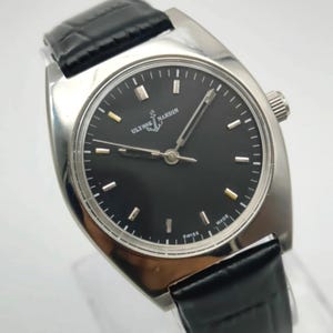 May include: A silver-toned wristwatch with a black face and a black leather band. The watch face features the brand name "Ulysse Nardin" and the word "Swiss" at the bottom. The watch has a rectangular shape with rounded edges.