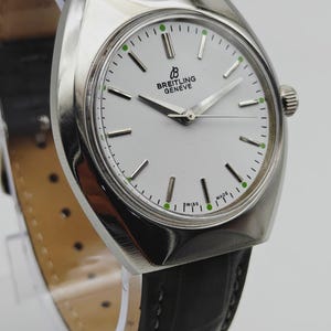 May include: A silver Breitling Geneve watch with a white face, black leather band, and green accents. The watch features a round face with the brand name and the words "SWISS MADE". The watch band is approximately 8 inches long.