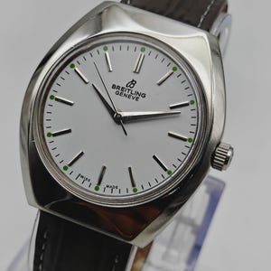 May include: A silver Breitling Geneve watch with a white face and black hands. The watch has a black leather band and green accents on the face. The watch face reads "SWISS MADE".