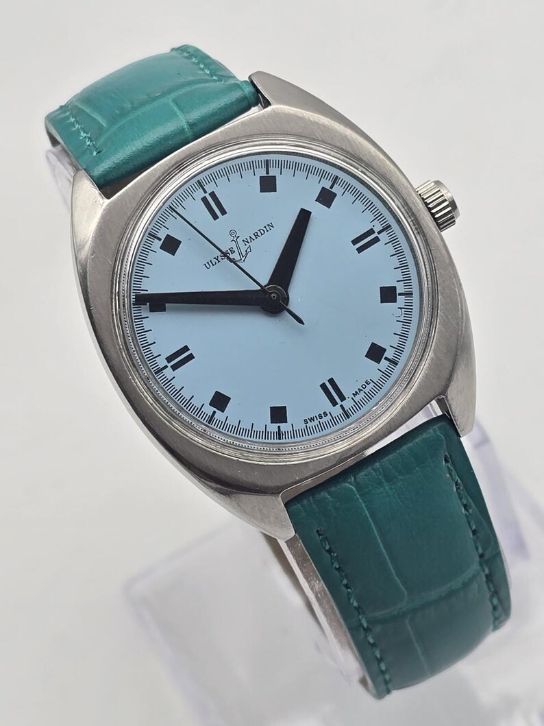 Pu&ograve; includere: Un orologio da polso vintage Ulysse Nardin con quadrante azzurro e lancette e indici neri. L'orologio ha una cassa argentata e un cinturino in pelle verde acqua. Sul quadrante sono presenti le scritte "Ulysse Nardin" e "Swiss Made".