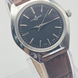 May include: A silver-toned Ulysse Nardin wristwatch with a black face and brown leather band. The watch face features silver hour markers and hands. The brand name and anchor logo are visible on the watch face. The watch is marked "Swiss Made".