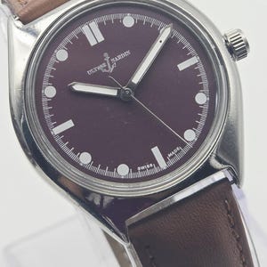 May include: A stainless steel wristwatch with a burgundy dial and a brown leather strap. The watch face features white hour markers and hands, with the brand name "Ulysse Nardin" visible. The watch has a classic, minimalist design.