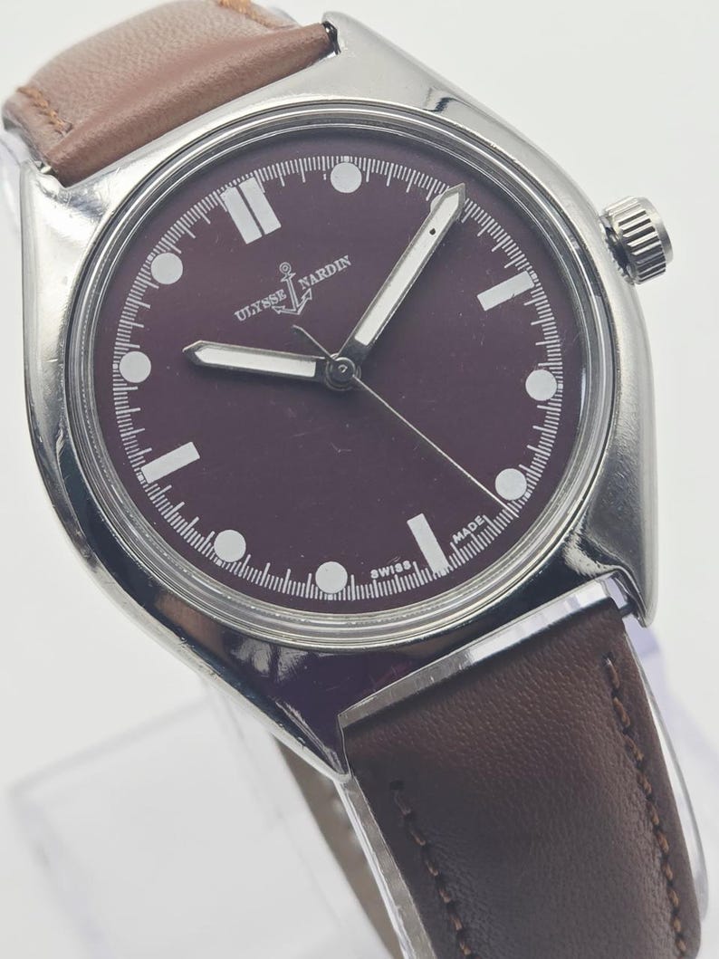 May include: A stainless steel wristwatch with a burgundy dial and a brown leather strap. The watch face features white hour markers and hands, with the brand name "Ulysse Nardin" visible. The watch has a classic, minimalist design.