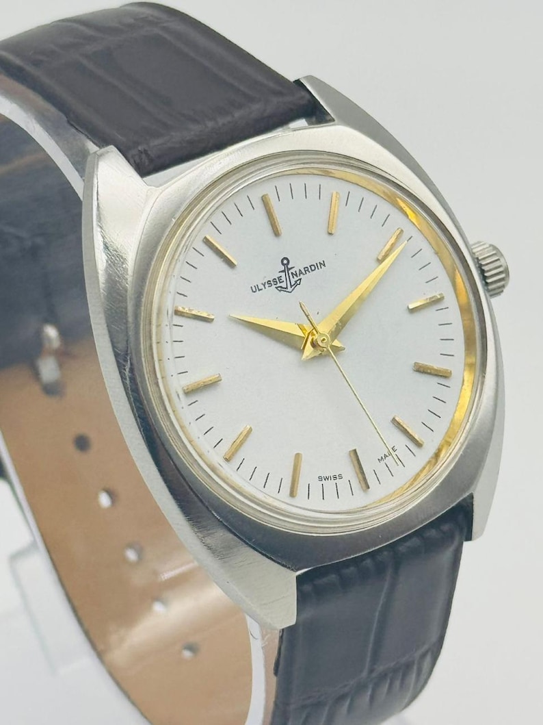 May include: A silver-toned Ulysse Nardin wristwatch with a white face, gold hands, and a black leather band. The watch face features the brand name and a small anchor logo. The watch is marked "Swiss Made".