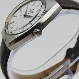 May include: A silver-toned wristwatch with a white face and a brown leather band. The watch face features the brand name "Breitling Geneve". The watch has a rectangular case and a crown on the side. The watch band is dark brown.