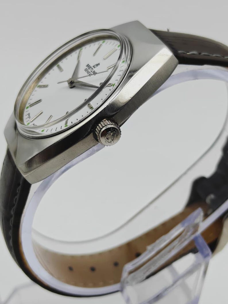 May include: A silver-toned wristwatch with a white face and a brown leather band. The watch face features the brand name "Breitling Geneve". The watch has a rectangular case and a crown on the side. The watch band is dark brown.
