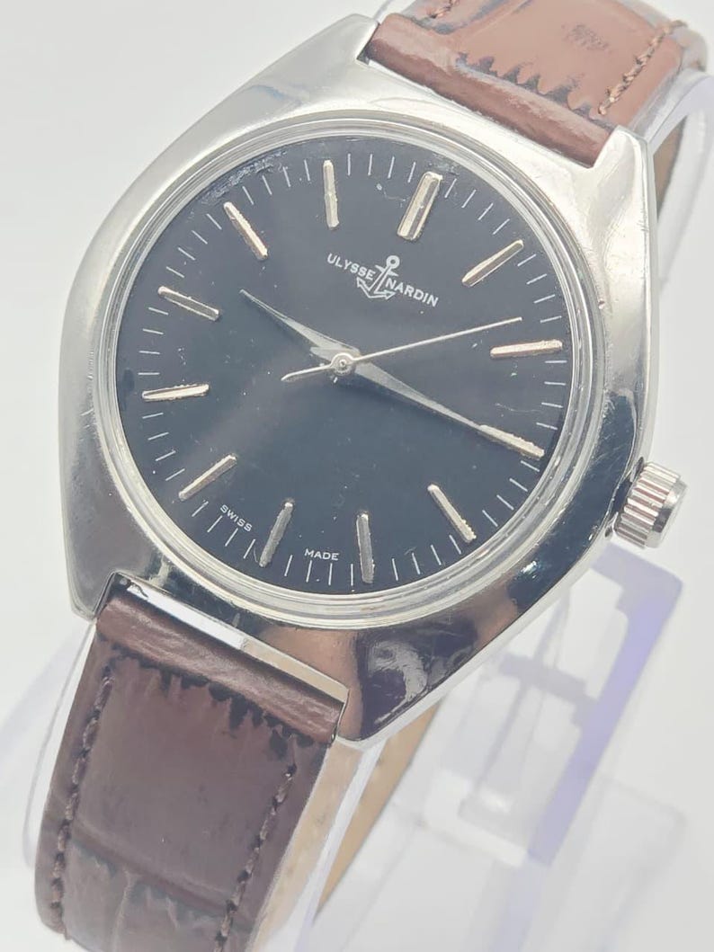 May include: A vintage Ulysse Nardin wristwatch with a black dial and silver-tone hands and markers. The watch features a stainless steel case and a brown leather strap. The dial includes the brand name and the words "Swiss Made".