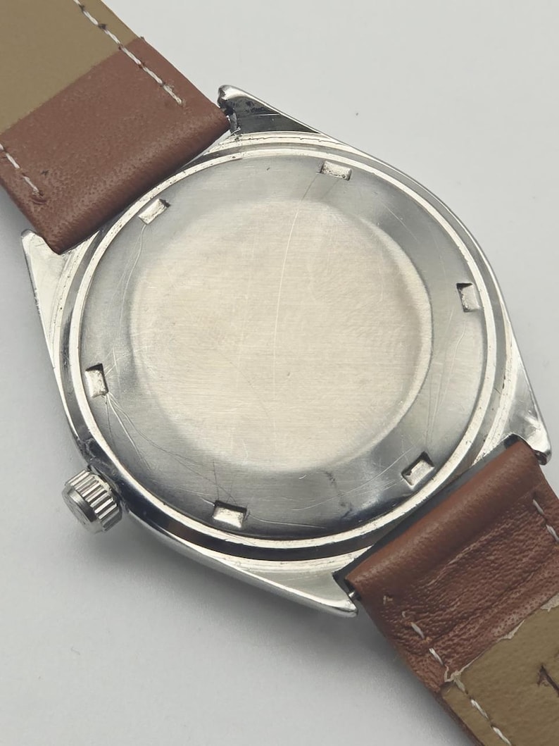 May include: Close-up of the back of a vintage wristwatch. The watch has a silver-colored case with a round, brushed metal back. Brown leather watch straps are attached. The watch's crown is visible on the side.