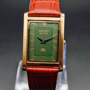Puede incluir: Un reloj Seiko Quartz dorado con una esfera rectangular verde y n&uacute;meros romanos. La correa es de cuero marr&oacute;n. La esfera del reloj tiene el texto "SEIKO QUARTZ".