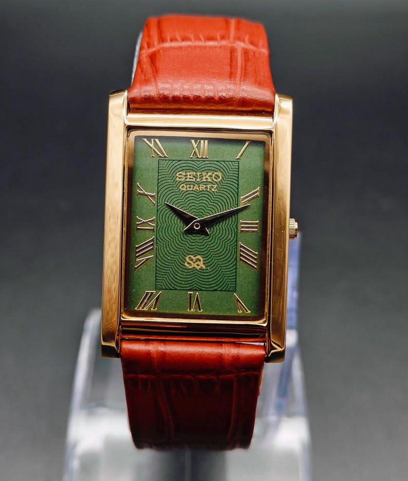 Puede incluir: Un reloj Seiko Quartz dorado con una esfera rectangular verde y n&uacute;meros romanos. La correa es de cuero marr&oacute;n. La esfera del reloj tiene el texto "SEIKO QUARTZ".