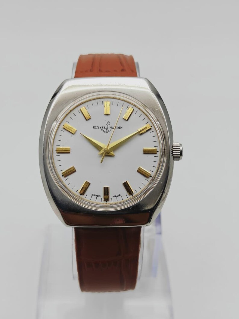 K&ouml;nnte beinhalten: Eine Vintage-Armbanduhr von Ulysse Nardin mit silberfarbenem Geh&auml;use und wei&szlig;em Zifferblatt. Die Uhr hat goldfarbene Zeiger und Markierungen, ein braunes Lederarmband und den Text "Ulysse Nardin". Die Uhr ist mit "Swiss Made" gekennzeichnet.