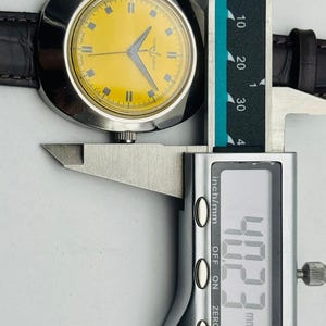 May include: A vintage wristwatch with a yellow face and silver case is measured with a digital caliper. The watch has a brown leather band. The caliper reads 40.23 mm, which is approximately 1.58 inches.