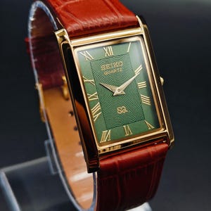 Puede incluir: Un reloj Seiko Quartz rectangular dorado con esfera verde y n&uacute;meros romanos. El reloj tiene una correa de cuero marr&oacute;n. La esfera del reloj tiene el logotipo de Seiko Quartz y un patr&oacute;n texturizado. El reloj se exhibe sobre un soporte transparente.