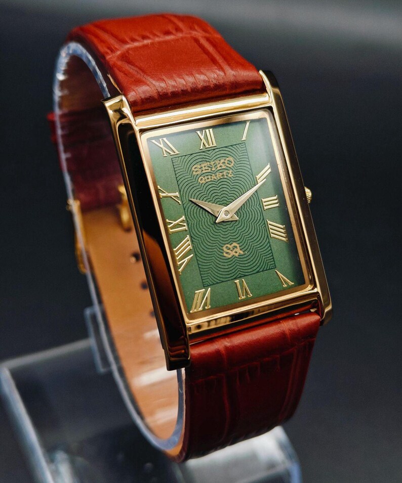 Puede incluir: Un reloj Seiko Quartz rectangular dorado con esfera verde y n&uacute;meros romanos. El reloj tiene una correa de cuero marr&oacute;n. La esfera del reloj tiene el logotipo de Seiko Quartz y un patr&oacute;n texturizado. El reloj se exhibe sobre un soporte transparente.