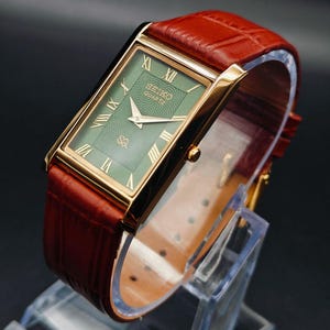 Puede incluir: Un reloj Seiko Quartz rectangular dorado con esfera verde y n&uacute;meros romanos. El reloj tiene una correa de cuero marr&oacute;n. El reloj se exhibe sobre un soporte de pl&aacute;stico transparente sobre un fondo oscuro.