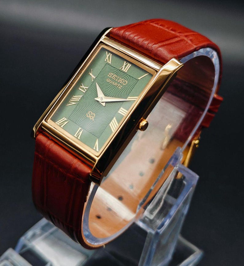 Puede incluir: Un reloj Seiko Quartz rectangular dorado con esfera verde y n&uacute;meros romanos. El reloj tiene una correa de cuero marr&oacute;n. El reloj se exhibe sobre un soporte de pl&aacute;stico transparente sobre un fondo oscuro.