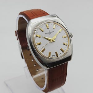 K&ouml;nnte beinhalten: Eine Vintage-Armbanduhr von Ulysse Nardin mit silberfarbenem Geh&auml;use und wei&szlig;em Zifferblatt mit goldfarbenen Zeigern und Markierungen. Die Uhr hat ein braunes Lederarmband. Das Zifferblatt zeigt den Markennamen und die Worte "SWISS MADE".