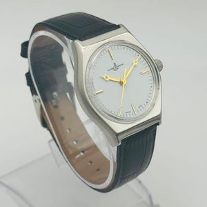 May include: A silver-toned wristwatch with a white face and gold hands and markers. The watch has a black leather band. The watch face has the words "ULYSSSE NARDIN" printed on it.