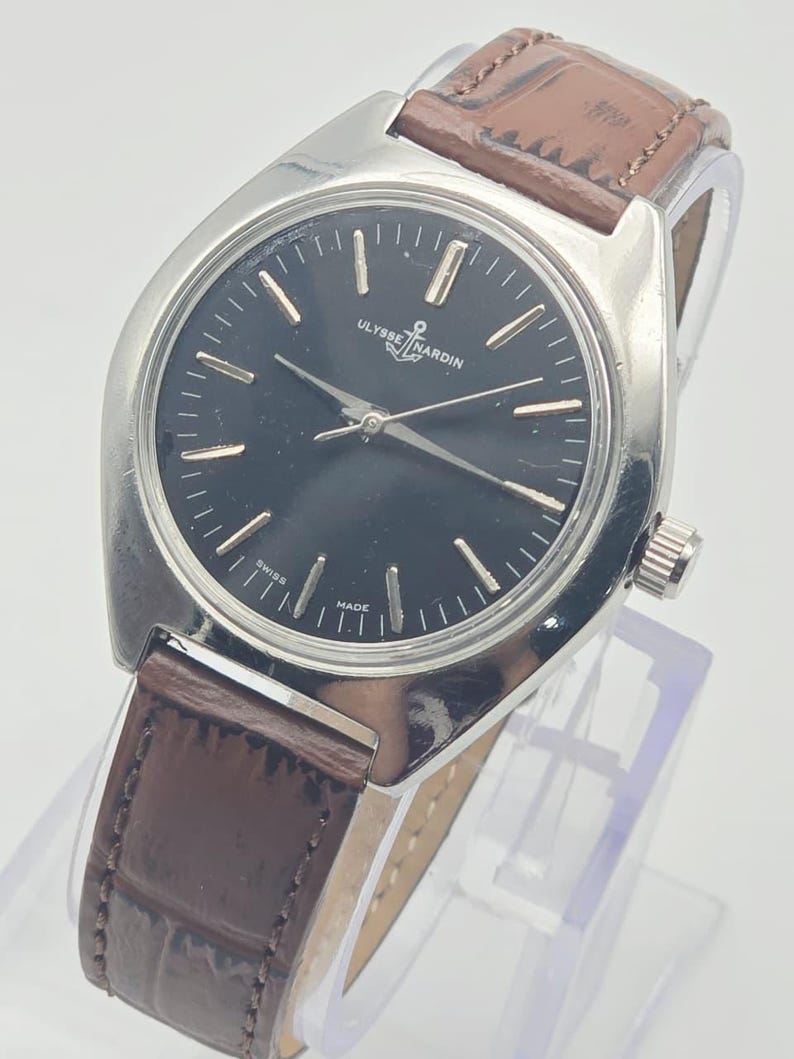 May include: A silver-toned Ulysse Nardin wristwatch with a black dial and brown leather band. The watch face has silver hour markers and hands. The band is a dark brown with a textured pattern.
