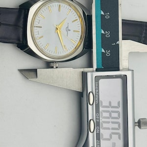 May include: A silver-toned wristwatch with a white face, gold hands, and a black leather band. The watch is being measured with a digital caliper, showing a width of 38.05 mm.