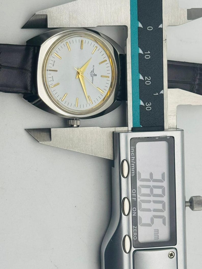 May include: A silver-toned wristwatch with a white face, gold hands, and a black leather band. The watch is being measured with a digital caliper, showing a width of 38.05 mm.