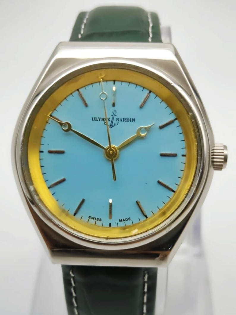 May include: A silver Ulysse Nardin wristwatch with a light blue face and gold accents. The watch features gold hands and a green leather strap. The words "SWISS MADE" are printed on the watch face.