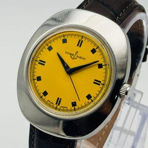 May include: A vintage Ulysse Nardin wristwatch with a bright yellow face and black hands and markers. The watch has a silver-tone case and a dark brown leather band. The watch face displays the text "Ulysse Nardin" and "Swiss Made".