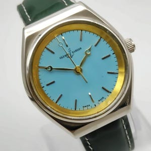 May include: A silver Ulysse Nardin watch with a light blue face and gold accents. The watch has a green leather strap and is marked "Swiss Made". The watch hands are gold-coloured.