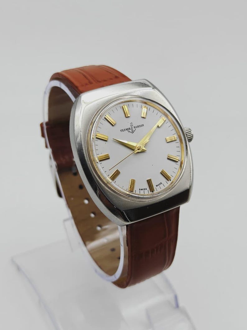 K&ouml;nnte beinhalten: Eine Vintage-Armbanduhr von Ulysse Nardin mit silberfarbenem Geh&auml;use und wei&szlig;em Zifferblatt mit goldfarbenen Zeigern und Markierungen. Die Uhr hat ein braunes Lederarmband. Das Zifferblatt zeigt den Markennamen und die Aufschrift "SWISS MADE".