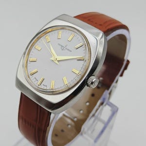 K&ouml;nnte beinhalten: Eine Vintage-Ulysse Nardin Uhr mit einem silberfarbenen quadratischen Geh&auml;use und einem wei&szlig;en Zifferblatt. Die Uhr hat goldfarbene Zeiger und Markierungen sowie ein braunes Lederarmband. Die Uhr wird auf einem transparenten St&auml;nder ausgestellt.
