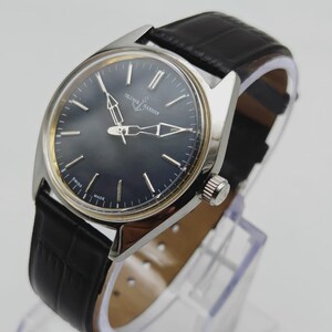Puede incluir: Un reloj de pulsera Ulysse Nardin plateado con esfera azul oscuro y correa de cuero negro. El reloj tiene agujas e indicadores de hora plateados. La correa tiene un patr&oacute;n de cocodrilo. El reloj se exhibe sobre un soporte transparente.