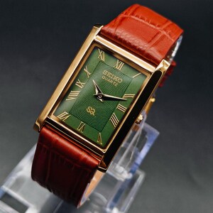 Puede incluir: Un reloj Seiko Quartz rectangular con caja dorada y esfera verde con n&uacute;meros romanos. El reloj tiene una correa de cuero marr&oacute;n. El reloj se exhibe sobre un soporte transparente.