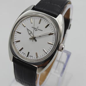 May include: A silver wristwatch with a white face and a black leather strap. The watch face has black hour markers and the brand name "ULYSSSE NARDIN". The watch is displayed on a clear stand.