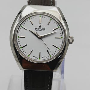 May include: A silver Breitling Geneve watch with a white face, black hands, and a brown leather strap. The watch face has green accents and the words "SWISS MADE". The watch is displayed on a clear stand.