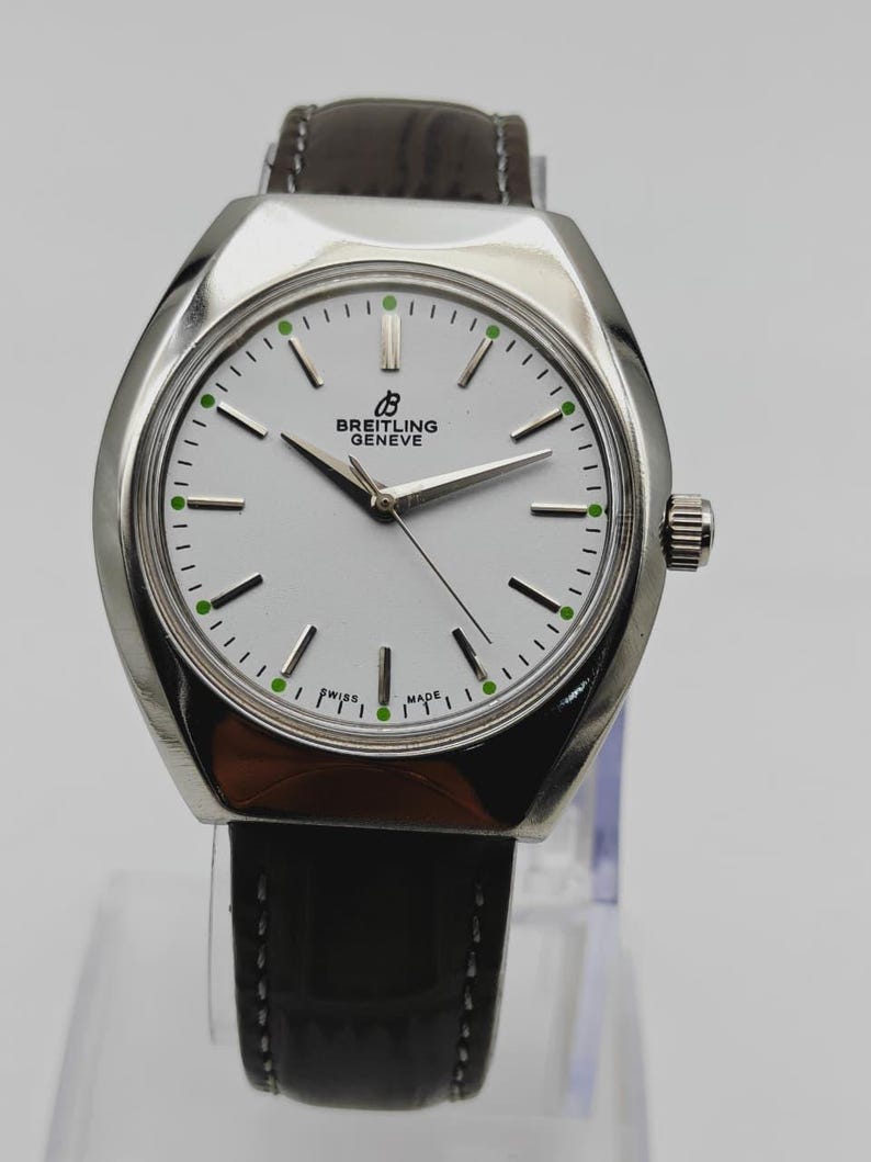 May include: A silver Breitling Geneve watch with a white face, black hands, and a brown leather strap. The watch face has green accents and the words "SWISS MADE". The watch is displayed on a clear stand.